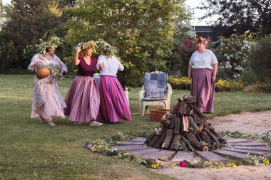 Yaz gündönümünün akşamında kafalarında çelenk ve renkli giysiler olan kadınlar kamp ateşinin etrafında dans ederler. Dışarıda, bahçede..