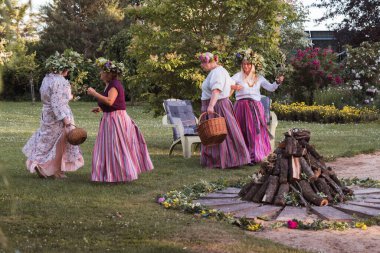 Yaz gündönümünün akşamında kafalarında çelenk ve renkli giysiler olan kadınlar kamp ateşinin etrafında dans ederler. Dışarıda, bahçede..