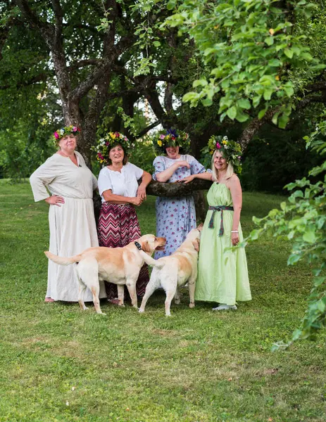 Ev yapımı çelenklerle süslenmiş dört kadın, bir yaz gündönümü akşamında, yanlarında bir Labrador ile bahçede dikiliyorlar..