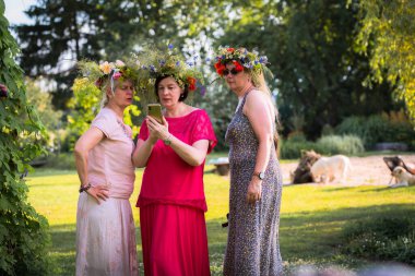 Başlarında yaz gündönümü çelenkleri olan üç kadın, yan yana durup aynı telefona bakıyorlar; ortadaki, kırmızı elbiseli, telefonda ne olduğunu gösteriyor.