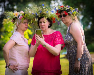 Başlarında yaz gündönümü çelenkleri olan üç kadın, yan yana durup aynı telefona bakıyorlar; ortadaki, kırmızı elbiseli, telefonda ne olduğunu gösteriyor.