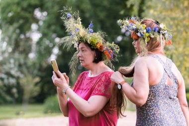 Başlarında yaz gündönümü çelengi olan iki kadın yan yana durur ve kırmızı elbiseli olanın tuttuğu telefona bakarlar..