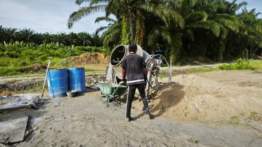 Perak, Malezya. 30 Mayıs 2025: Taman Indah Permai şantiyesindeki kum ve çimento torbalarının yanında çimento karıştırma makinesinde çalışan bir işçi.