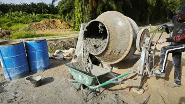 Perak, Malezya. 30 Mayıs 2025: Taman Indah Permai şantiyesindeki kum ve çimento torbalarının yanında çimento karıştırma makinesinde çalışan bir işçi.