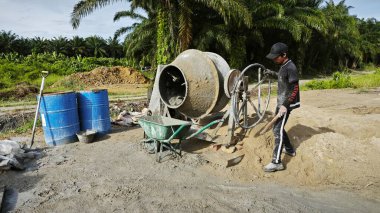 Perak, Malezya. 30 Mayıs 2025: Taman Indah Permai şantiyesindeki kum ve çimento torbalarının yanında çimento karıştırma makinesinde çalışan bir işçi.