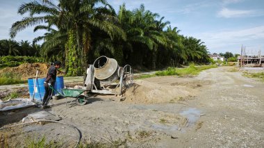 Perak, Malezya. 30 Mayıs 2025: Taman Indah Permai şantiyesindeki kum ve çimento torbalarının yanında çimento karıştırma makinesinde çalışan bir işçi.