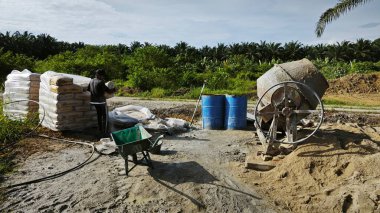 Perak, Malezya. 30 Mayıs 2025: Taman Indah Permai şantiyesindeki kum ve çimento torbalarının yanında çimento karıştırma makinesinde çalışan bir işçi.