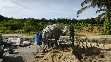 Perak, Malezya. 30 Mayıs 2025: Taman Indah Permai şantiyesindeki kum ve çimento torbalarının yanında çimento karıştırma makinesinde çalışan bir işçi.