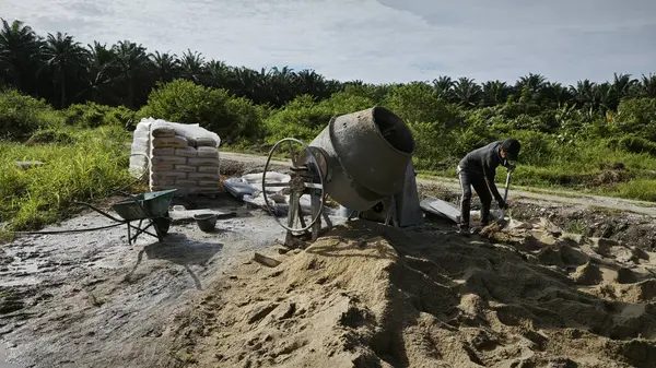 Perak, Malezya. 30 Mayıs 2025: Taman Indah Permai şantiyesindeki kum ve çimento torbalarının yanında çimento karıştırma makinesinde çalışan bir işçi.