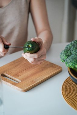 Genç bir kadın, ahşap kesme tahtasından avokado kesiyor. Sağlıklı vejetaryen yemeği. Sağlıklı yemek pişirmek. Taze yeşil avokado.