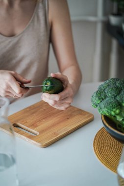 Genç bir kadın, ahşap kesme tahtasından avokado kesiyor. Sağlıklı vejetaryen yemeği. Sağlıklı yemek pişirmek. Taze yeşil avokado.