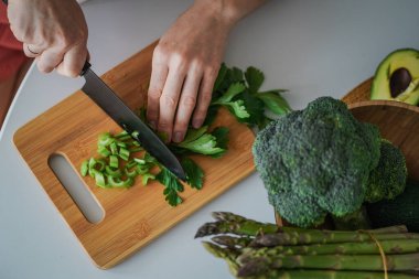 Tahta kesme tahtasındaki kereviz kesme işlemi. Sağlıklı yemek pişirmek. Parlak yeşil kereviz, brokoli, kuşkonmaz ve avokado yakın plan.