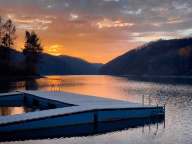 Lake surrounded by mountains reflecting in the water and a wooden pontoon at sunset