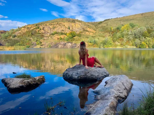 Woman wearing red dress and red flower in her hair  sitting on a rock near a lake surrounded by mountains and admiring the beautiful scenery around her