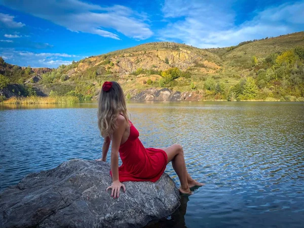 Woman wearing red dress and red flower in her hair  sitting on a rock near a lake surrounded by mountains and admiring the beautiful scenery around her