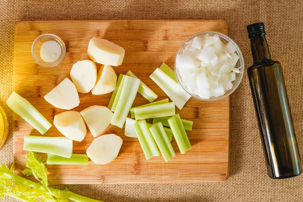 Raw vegetables, such as celery sticks, potatoes, chopped onion, olive oil, salt, and broth close-up on the kitchen table. Ingredients for a celery soup recipe