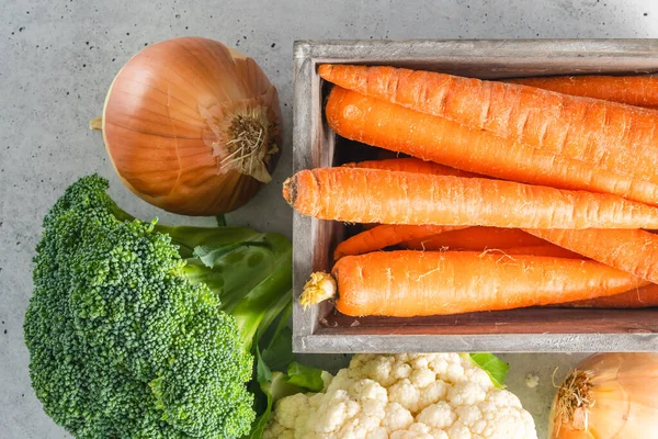 Carrots in a wooden box, broccoli, cauliflower, and onion close-up on a grey stone background, flat lay with copy space