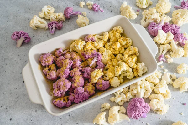 Cauliflower, white and purple, in baking pan with garlic seasoning, ready to be baked. Close-up view, grey stone background