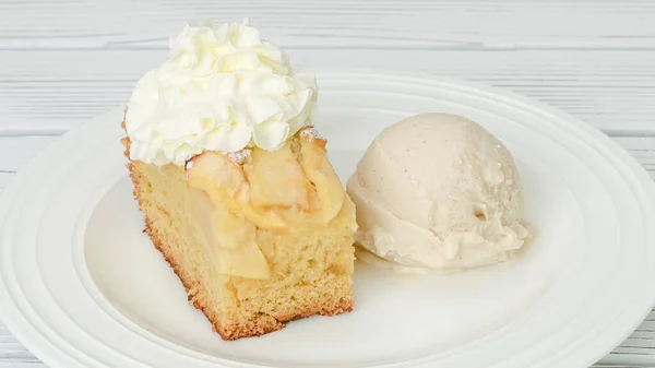 Slice of fresh baked apple cake with biscuit base and sliced apples and whipped cream topping served with vanilla ice cream close up on a plate on white background