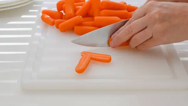 Woman cuts carrot. Fresh organic peeled baby carrots close up on cutting board on white kitchen table.