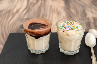 Beyaz çikolatalı vanilyalı puding, koyu çikolata soslu ve gökkuşağı parçalı, ahşap arka planda yakın plan.