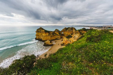 Kayalık oluşumları olan engebeli kıyı şeridi, kumlu bir plaj ve dramatik bulutlu bir gökyüzü, Algarve, Portekiz