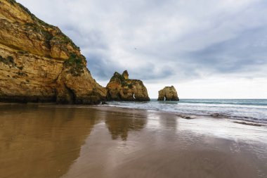 Çalkantılı okyanus dalgalarının dramatik bulutlu bir gökyüzünün altında kayalık kıyılarla buluştuğu engebeli kıyı şeridi doğal kemerler ve mağaralarla süslenerek büyüleyici ve atmosferik bir deniz manzarası yaratır.