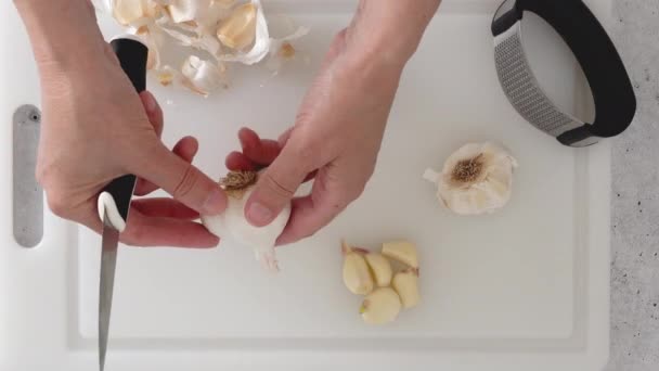 女性はキッチンテーブルの上に白いまな板の上にニンニクプレスツールを使用して新鮮なニンニクを押す — ストック動画
