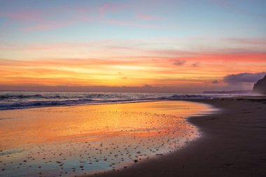 Sersemletici altın gün batımı kumlu sahilleri süslüyor, sıcak renklerle gökyüzünü boyuyor ve sakin plajda büyüleyici bir parıltı yayıyor.