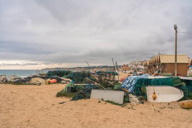 Fishing gear and boats scattered on a sandy beach under a cloudy sky. Algarve, Portugal, January 3, 2024 clipart