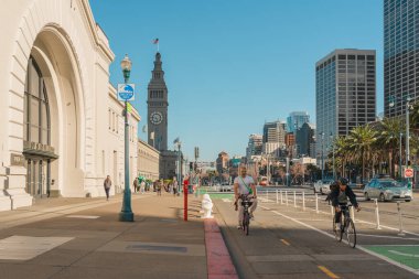 San Francisco, Kaliforniya, 8 Nisan 2024. The Embarcadero 'daki sokak sahnesinde bisikletçiler ve yayalar yüksek binalar ve palmiye ağaçlarıyla çevrili..