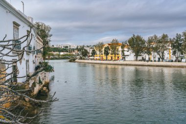 Tavira, Portekiz, 8 Ocak 2024. Portekiz 'in Tavira kentindeki nehir kıyısı manzarasında beyazlatılmış binalar, yapraklı ağaçlar ve sakin sulardaki yansımalar yer alıyor..