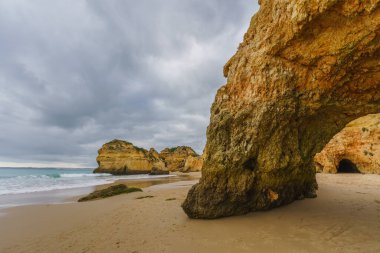 Engebeli kıyı şeridi kayalık oluşumlar, kumlu bir sahil ve dramatik bulutlu bir gökyüzü.
