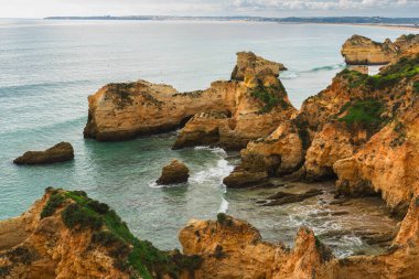 Mağaraları olan engebeli kıyı şeridi, çarpan dalgalar, kumlu plajlar ve arka planda bulutlu bir gökyüzü. Algarve, Portekiz