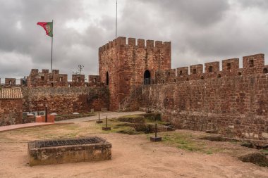 Silves Kalesi 'nin sağlam kırmızı tuğlalı surlarında bulutlu bir gökyüzüne karşı Portekiz bayrağı dalgalanıyor. Silves, Portekiz, 3 Ocak 2024.