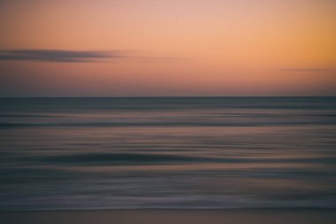 Bulanık okyanus dalgaları alacakaranlıkta yumuşak turuncu ve mavi gökyüzünün altında.