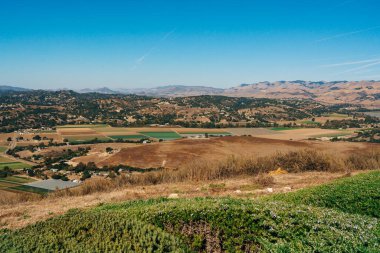 Hava manzarası, tarımsal tarlalarla süslenmiş dalgalı tepeleri ve berrak mavi gökyüzünün altındaki tuhaf evlerin siluetini gözler önüne seriyor..