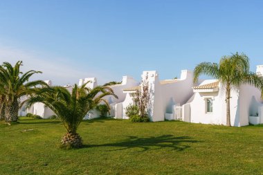 Palmiye ağaçlarıyla çevrili canlı turistik bir tatil beldesinde lüks tatilleri simgeleyen bir dizi apartman dairesi. Parlak güneşli bir gün, açık mavi bir gökyüzü.