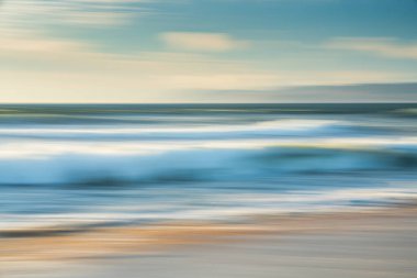 Hafif, havadar, soyut deniz manzarası. Geniş kumsal, deniz dalgaları, dağların silueti ve arka planda bulutlu gökyüzü, hareket bulanıklığı.