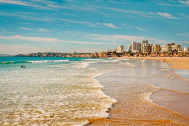 Nazik dalgalar Praia da Rocha 'nın altın kumları üzerinde yuvarlanıyor, arka planda şehir binaları ve uçurumlar var..