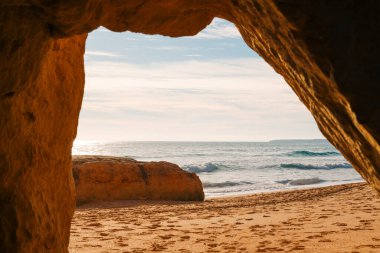 Güneşli bir gökyüzünün altında turkuaz okyanustan yükselen iki izole kaya formasyonuyla kumlu sahilde dalgalar yavaşça kırılır..