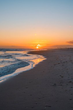 Sakin bir plajın üzerinde canlı bir günbatımı hafif dalgalar ve suyun kenarında yalnız bir figür..