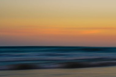 Bulanık okyanus dalgaları pastel bir günbatımı gökyüzüne doğru uzanıyor.