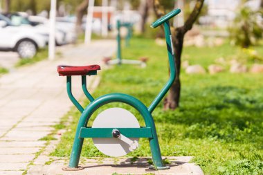 Açık havada yeşil metal çerçeveli ve kırmızı koltuklu staj bisikleti halka açık bir parka monte edilmiştir..