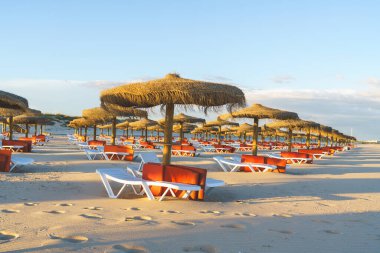 Gün batımında plajda şezlonglar ve saman şemsiyeler.