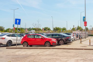 Loule, Portekiz- 27 Nisan 2025. Karışık arabalar, kırmızı bir araba dahil, güneşli bir otoparkta Tesla Superchargers 'ın yanına park edilmiş..