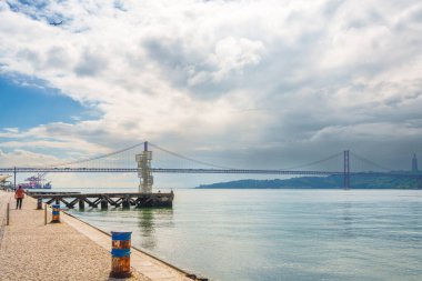 Lizbon, Portekiz 5 Mayıs 2025. 25 de Abril Köprüsü 'nün dramatik bir gökyüzünün altında nehir kenarında yürüyen bir yaya ile Tagus Nehri' ni kapladığı görüntüsü.