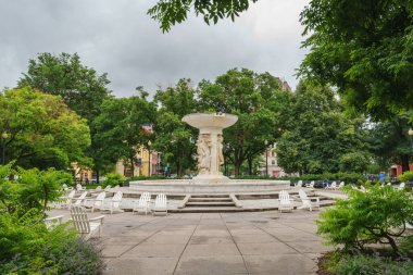 Washington D.C., ABD - 21 Mayıs 2025. Ağaçlarla dolu bir meydanda beyaz sandalyelerle çevrili klasik bir mermer çeşmenin geniş manzarası.
