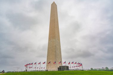 Washington D.C., ABD - 21 Mayıs 2025. Washington Anıtı, bulutlu bir günde, önü yeşil çimlerle kaplı Amerikan bayraklarıyla çevrili..