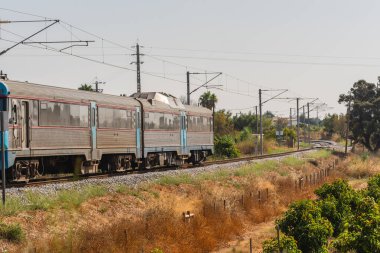 Bir yolcu treni, kurak bitki örtüsü ve kırsal alanla çevrili kıvrımlı bir elektrik hattı boyunca hareket eder..